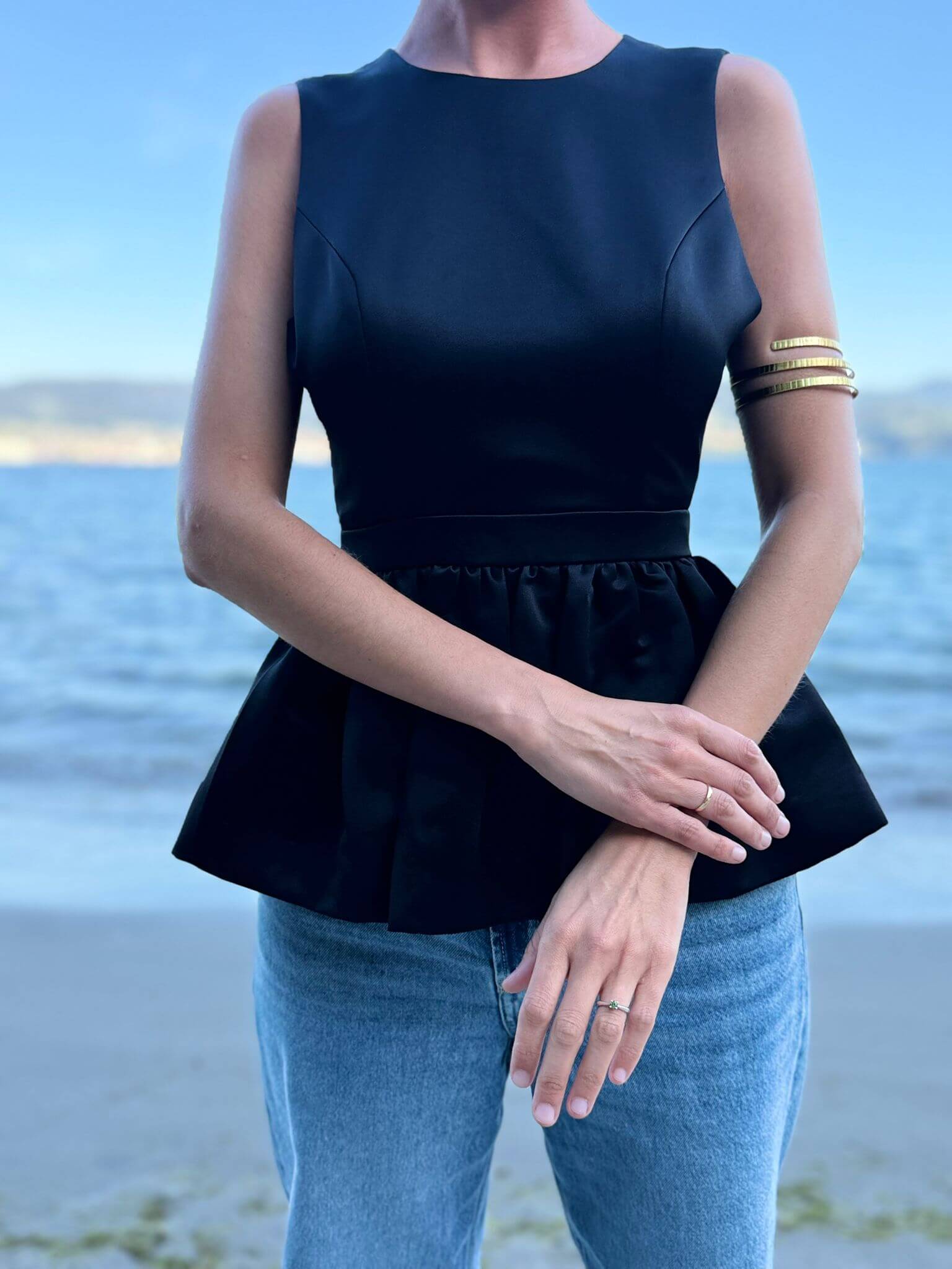Top negro de popelín con diseño entallado y peplum, frente a una playa con fondo marino.